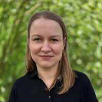 Portrait of a woman with shoulder-length light brown hair outdoors in a green park, wearing a dark collared shirt, smiling at the camera.