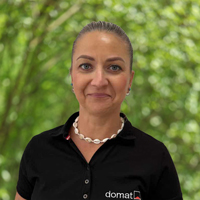Portrait of a woman with dark hair tied back, wearing a black domat polo, outdoors with green foliage behind her.