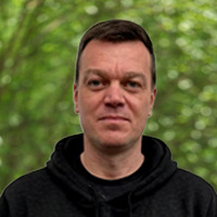 Portrait of a man with short dark hair wearing a black hoodie, standing in a green leafy park.