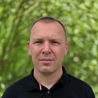 Portrait of a man outdoors in a green park, wearing a black polo shirt and looking at the camera.