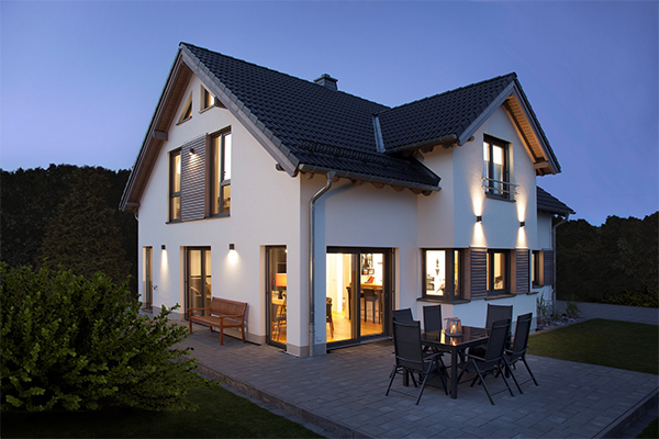 beleuchtetes, modernes Einfamilienhaus mit dunklem Dach in der Dämmerung. Es hat weiße Wände und mehrere Fenster, einige mit Rollläden. Eine Terrasse mit Gartenmöbeln ist vor dem Haus. Es könnte ein ISOWOODHAUS sein.