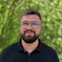 Portrait of a man with a beard and glasses, wearing a dark polo, outdoors with green trees in the background.