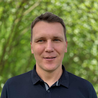 Smiling man in a dark polo shirt outdoors with green trees in the background.