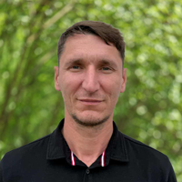 Portrait of a man with short brown hair wearing a black polo, standing outdoors with green foliage in the background.