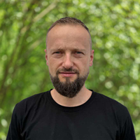 Head-and-shoulders portrait of a man with a short beard wearing a black shirt, standing among green trees.