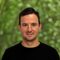 Portrait of a smiling man with short dark hair wearing a black shirt, outdoors with green foliage behind him.