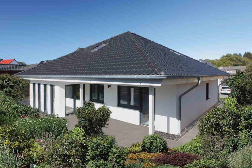 Ein weißer Bungalow mit dunklem Dach. Garten mit Pflanzen, gepflasterte Terrasse. Blauer Himmel.