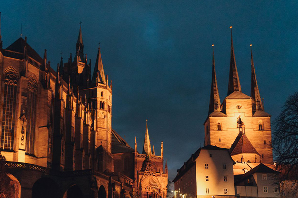 Night view of a Gothic cathedral with illuminated spires along a cobblestone area under a dark blue sky, warm lights highlighting the architecture on the left and right sides.