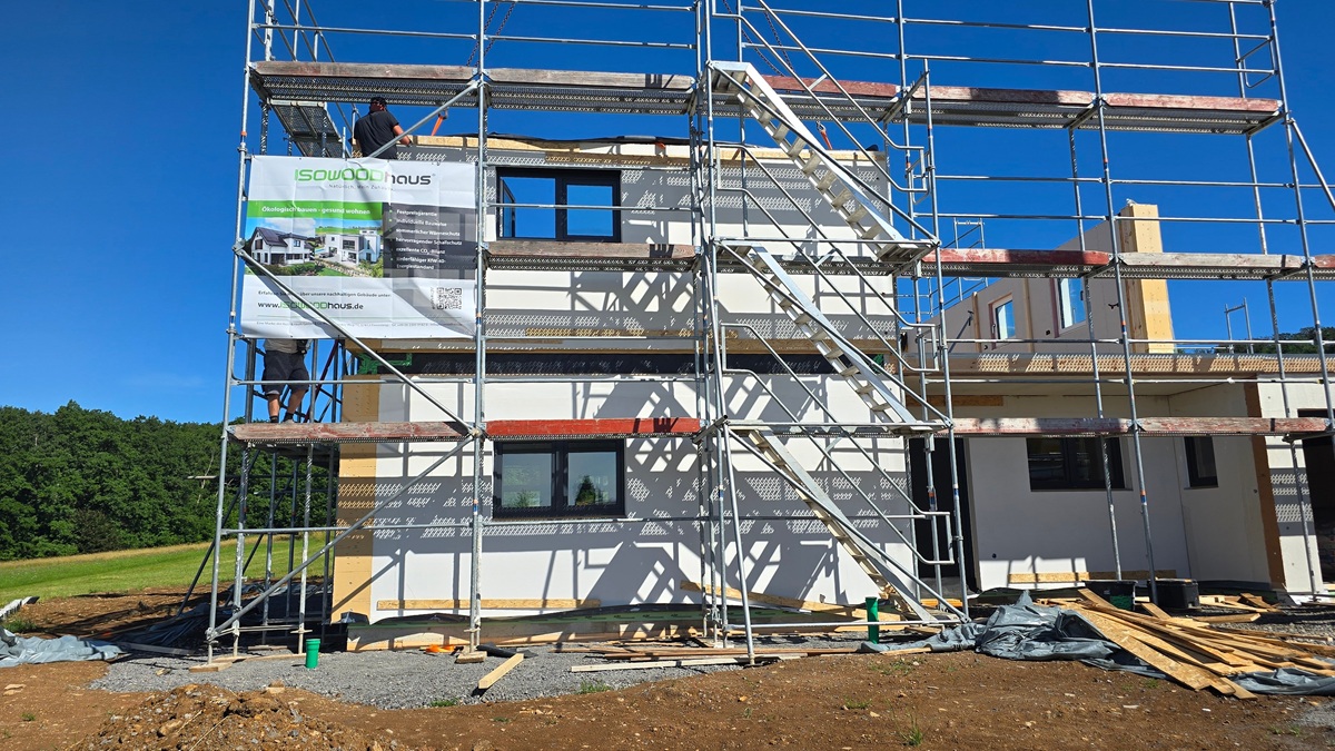 Two construction workers on metal scaffolding around a partially built, white-and-gray house under a bright blue sky with a banner hanging on the left side of the scaffold.