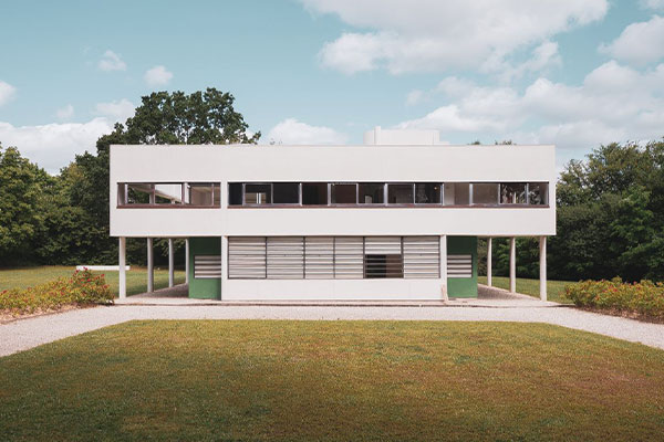 White modernist two-story building on stilts with long rectangular windows, set in a park with gravel paths and green lawn.