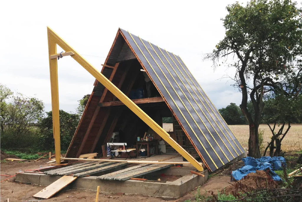 A-frame cabin under construction with yellow framing and roof panels, scattered tools outside in a rural setting.