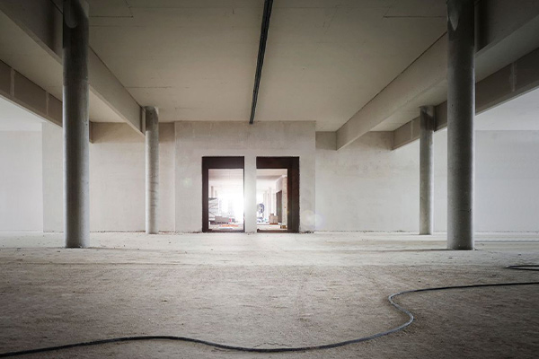 Empty concrete construction space with tall pillars and a central doorway framing bright light beyond; dusty floor with a loose cable in the foreground.