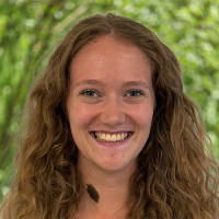 Young woman with long curly hair smiling outdoors with green trees behind her.