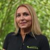 Portrait of a woman with shoulder-length blonde hair, wearing a black polo with a green logo, standing outdoors among green trees.
