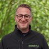 Portrait of a man with glasses wearing a black zip-up jacket with a green logo, outdoors in a leafy setting.