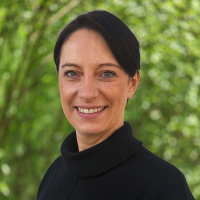 Smiling woman with short dark hair wearing a black turtleneck, outdoors with green foliage in the background.