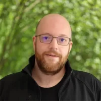 Eine Person in schwarzem Hemd vor grünem, unscharfem Hintergrund. Klare Hauptfokussierung.