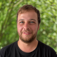 Close-up portrait of a smiling man with short light brown hair and a beard, wearing a black shirt, outdoors with green foliage in the background.
