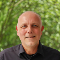Portrait of a smiling bald man in a black shirt outdoors with a blurred green foliage background