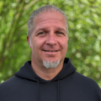 Smiling man with short gray hair and goatee wearing a dark hoodie outdoors, blurred green trees in the background